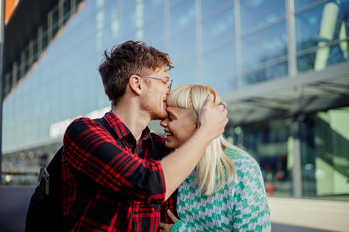 Portrait of a young couple sitting on a city street downtown and kissing. A trendy man is kissing his girlfriend in forehead while hugging her. An affectionate couple is having romantic moments.