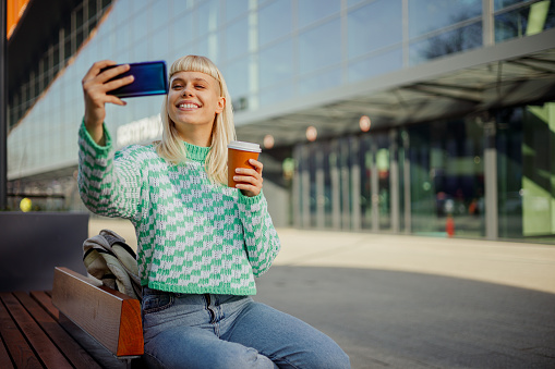 A trendy female influencer is sitting on a bench downtown and making selfies. An urban hipster girl is using her phone for taking selfies with coffee to go. A girl is photo messaging with mobile app.