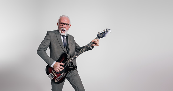Excited senior rock musician in suit singing with mouth open and playing guitar on white background