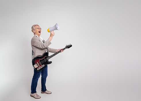 Excited senior businesswoman shouting over megaphone and playing electric guitar on white background