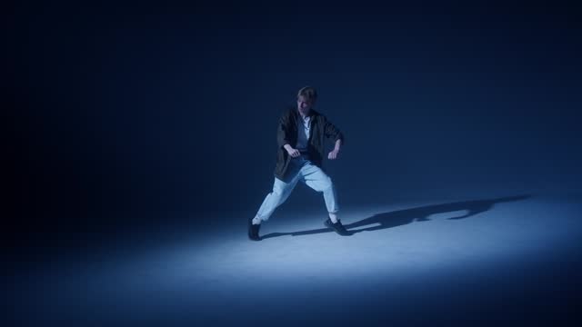 Young blonde guy dancing modern choreography in dark studio background. Talented choreographer man showing the new hip hop movements. Shooting a dance video clip. Hip hop school for teenagers.