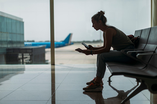 With concern, the young woman uses her phone to check her flight reservation while sitting at the airport bench