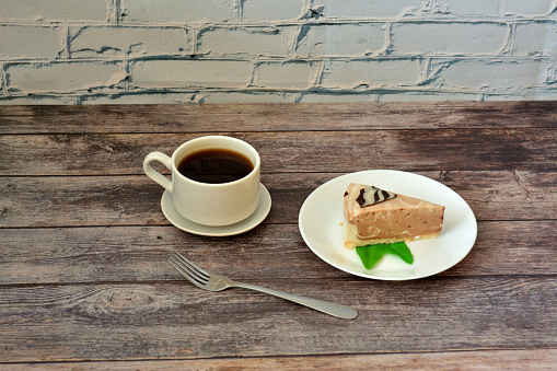 A plate with a slice of fresh cream cheesecake with mint and a cup of hot black coffee with a fork on a wooden table. Close-up.