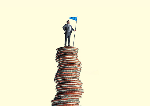 A man stand while holding a flag on top of a tall stack of coins in front of a yellow background.