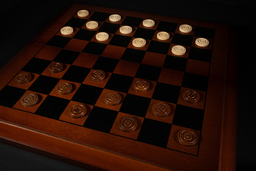 Close up shot of wooden checker pieces on a wooden checker board on a black background.
