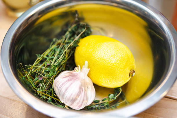 A stainless steel bowl containing fresh ingredients: a whole lemon, garlic bulb, and aromatic herbs, ready for culinary use.
