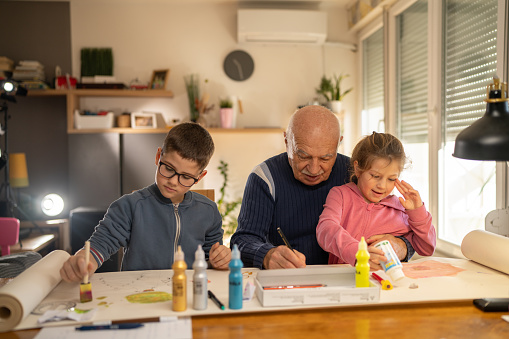 Happy grandfather sitting at the table and drawing with his grandchildren at home