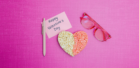 Pink background with white and pink rice treat heart, pink glasses, pen and Happy Valentines Day note.  Romantic