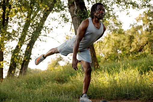 Young African man standing on one leg and stretching before jogging in nature