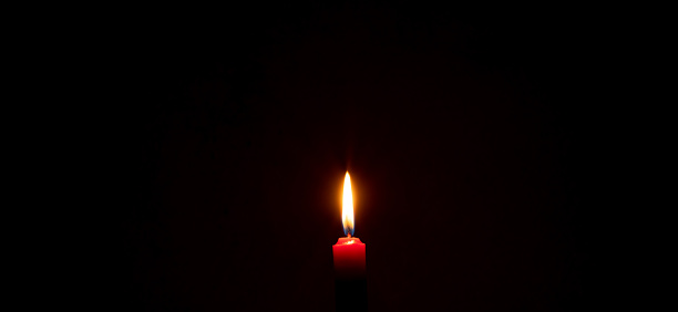 A single burning candle flame or light is glowing on a red candle on black or dark background on table in church for Christmas, funeral or memorial service with copy space