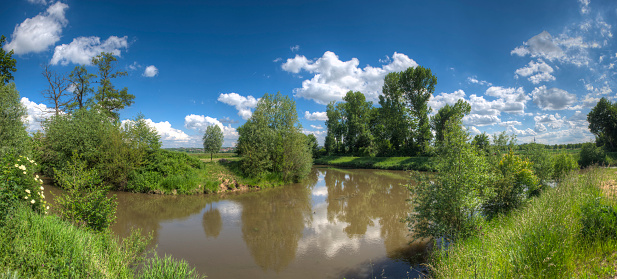 Panoramic view of a brown-colored river in a natural landscape with green riverbank vegetation, deciduous trees, fields and natural meadows in fine weather and with scattered clouds