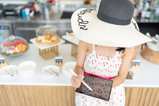 Top view of an Asian female tourist's hat Using a laptop to do business in front of the food department of a hotel restaurant, wearing a dress, with a menu on the back. Desserts and drinks