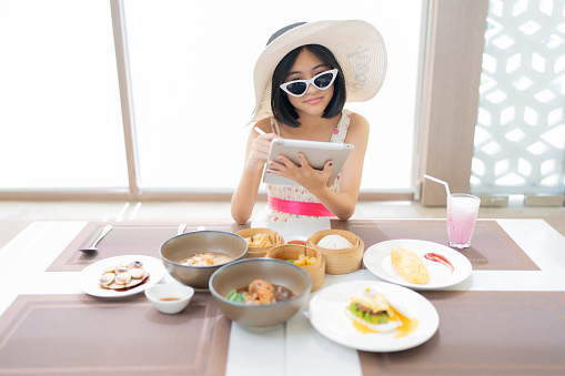 Asian female tourist relaxing on vacation at hotel Wear a hat and dress. Have breakfast On the table there are Chinese and European food, as well as desserts and drinks. Use a laptop to do business.