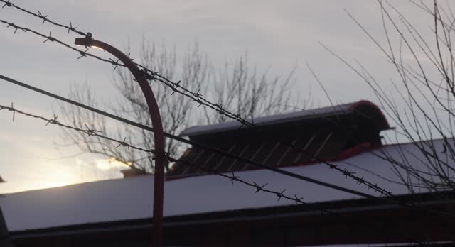 Barbed wire fence in the foreground, with a snow-covered building roof and bare tree branches against a cloudy sky in the background