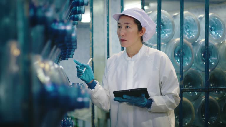 A female worker in lab attire examines water bottle production with a tablet in a factory. HACCP System in Bottled Water Production