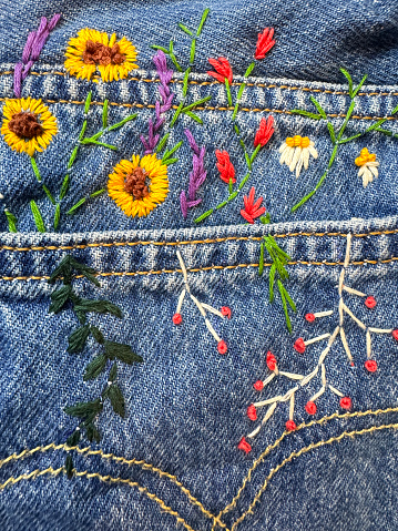 Stock photo showing close-up view of a pair of blue denim jeans sporting a customised design of embroidered flowers and leaves.