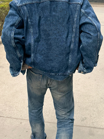 Stock photo showing close-up, rear view of an Indian man wearing a denim jacket and jeans adjusting trousers by pulling them up whilst walking through a paved area.