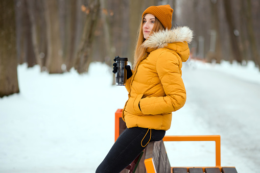 Young woman in yellow winter jacket drinking tea from the thermo cup near the bench in the winter park.
