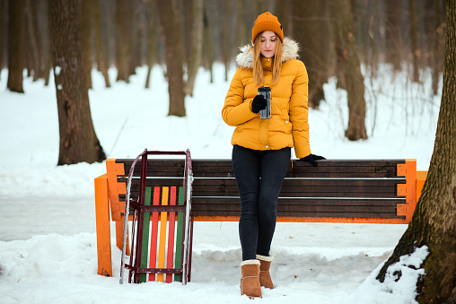 Beautiful young woman in warm winter clothes with sleigh drinking tea near the bench in the winter park.