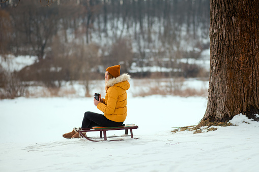 Young woman in yellow winter jacket sitting on a sleigh and drinking tea from the thermo cup in the winter park.