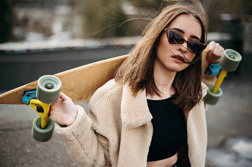 Young brunette with professional skateboard on shoulders spending time on roof of high building. Stylish woman wearing casual clothes and modern sunglasses.