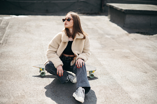 Cheerful beautiful woman in trendy clothes having fun while sitting on skateboard outdoors. Urban area with modern building on background. Lifestyles of youth people.