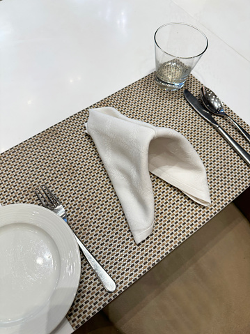 Stock photo showing close-up, elevated view of a restaurant table, which has been neatly laid and set for either breakfast, a lunchtime meal or an evening family dinner. On the dining table is a woven brown placemat, upon which stainless steel cutlery knife, fork and spoon have been laid, as well as a white side plate and an empty water glass, complete with cloth napkin.