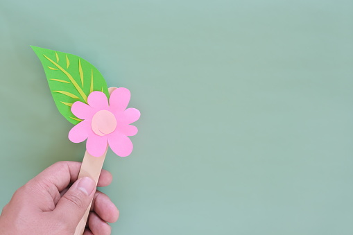 Human hand holding flower with leaf paper cutout design in green background with copy space. Spring season minimalist backdrop.