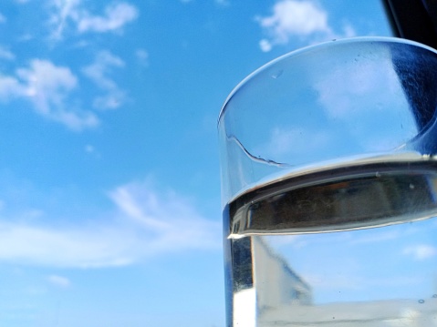 Refreshing purified water in transparent glass with reflection against blue sky, cloud and greeneries