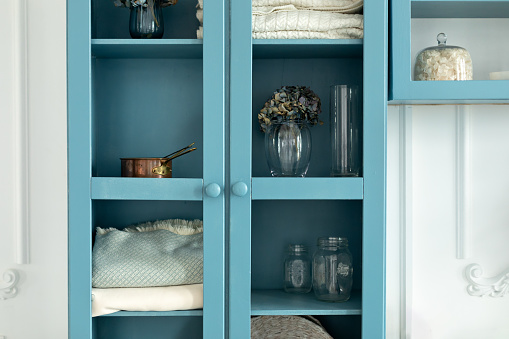 Wooden blue book case with decor and flowers in vase near wall in living room. Cozy home interior with decoration on shelves with utensils. Organization at home