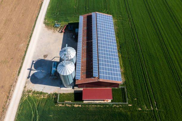 Aerial view of an agricultural building with solar panels on the roof. Two silos, agricultural equipment and dirt roads between green fields in spring are also visible.
