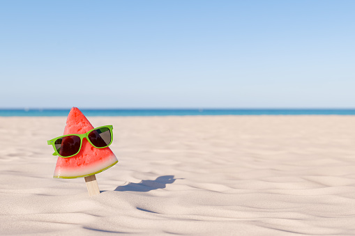 3d rendering of a whimsical watermelon slice on a stick sporting green sunglasses against a sunny beach backdrop with the ocean in the distance. Summer vacation concept.