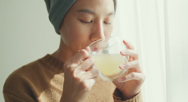 Closeup of young Asian woman sick with cancer in a headscarf enjoys the warmth of the morning sun and a hot drink by the window at home. Living with cancer.