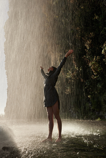 Young happy woman in raincoat having fun while standing under the waterfall in nature.