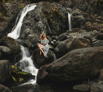 Young smiling woman day dreaming while sitting on a rock by the waterfall in nature.