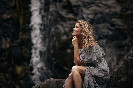 Smiling woman day dreaming while sitting on the rock by the waterfall in nature.