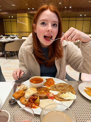 Stock photo showing close-up view of attractive, red haired, young woman in a self-service buffet restaurant eating masala dosa pancake with chutney, grilled tomato, a pile of baked beans, sausage and a medu vada (savoury doughnut).