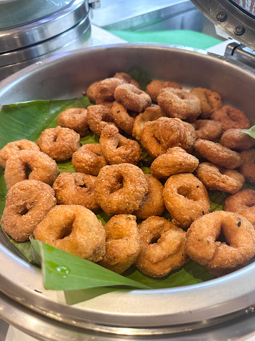 Stock photo showing close-up, elevated view of pile of medu vada (savoury doughnuts) being served in a banana leaf lined, stainless steel hot plate warmer dish with open lid at a restaurant self service buffet. Medu vada savoury doughnut fritters are made from Vigna mungo (black lentil) batter and are a popular breakfast snack.