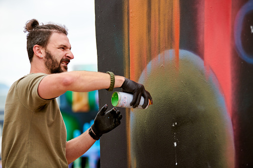 Attractive male artist is painting picture with paint spray can spraying it onto canvas at outdoor street exhibition, side view of man art maker
