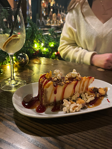 Stock photo showing close-up view of a slice of caramel cheesecake with digestive biscuit base, decorated with honeycomb pieces and popcorn kernels, and drizzled with chocolate sauce, in a restaurant setting.