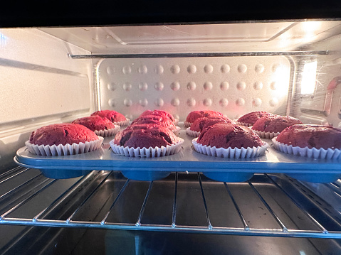 Stock photo showing close-up view of a shelf inside of a hot oven with freshly baked rows of homemade, red velvet cupcakes in paper cake cases rising in a turquoise non-stick baking tray. Home baking concept.