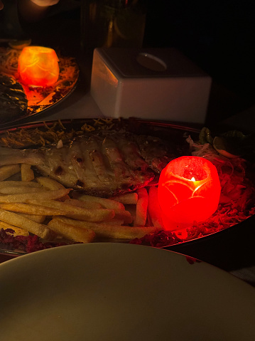 Stock photo showing close-up, night-time view of a romantic, beach restaurant, alfresco dining table backlit by a candle flame in a glass hurricane lamp. This romantic meal for two features battered fish and chips on a large plate, with salad garnish and an illuminated candle carved from a tomato.