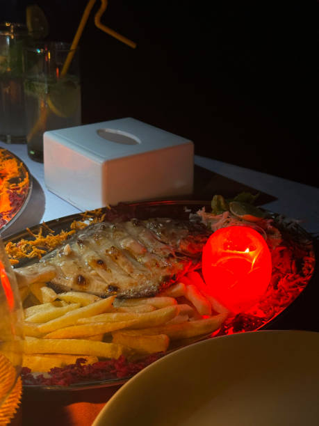 istock Image of backlit dinner plate containing meal of battered fish and chips served with side salad and candle made of carved tomato, napkin dispenser box, glasses of mojito cocktails, al fresco beach restaurant at night, focus on foreground 1979872590