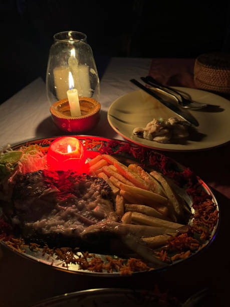 Image of backlit dinner plate containing meal of battered fish and chips served with side salad and candle made of carved tomato, glass hurricane lamp candle holder, al fresco beach restaurant at night, focus on foreground