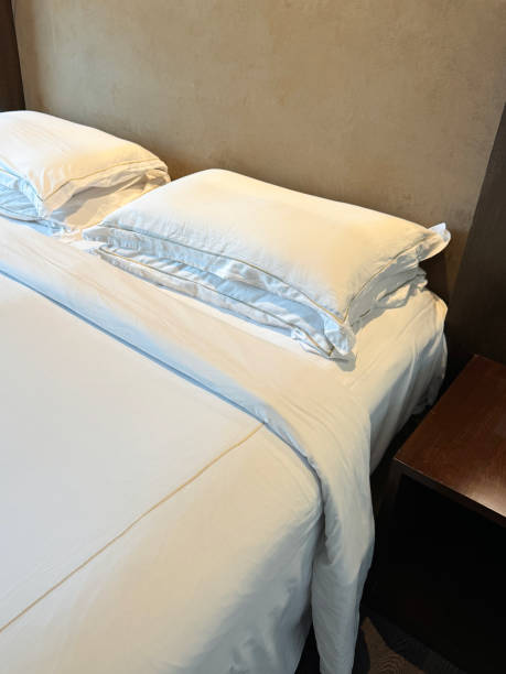 istock Close-up image of white duvet bed covered double bed, stack of white hotel pillows, folded bedding, suede effect headboard, nightstand, focus on foreground, elevated view 1979921486