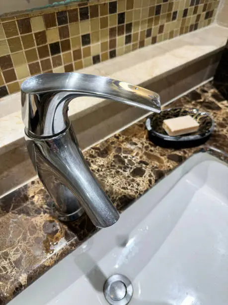 Stock photo showing close-up view of chrome, mixer tap on white ceramic sink in bathroom with brown marble countertop.