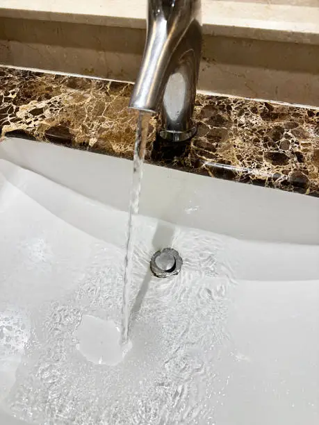 Stock photo showing close-up view of chrome, mixer tap on white ceramic sink in bathroom with brown marble countertop.