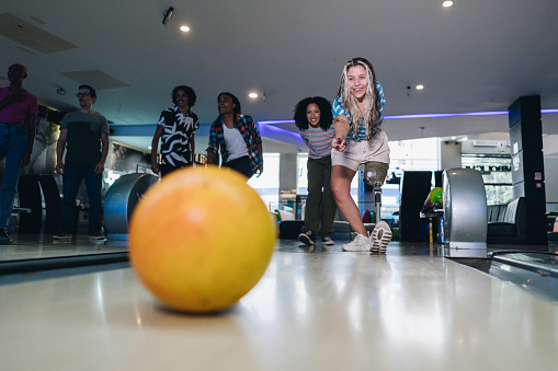 Young woman with a prosthesis leg throwing a bowling ball at a bowling club with friends