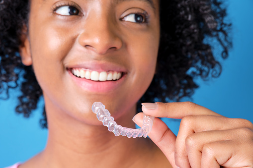 closeup of young colombian woman with afro hair smiling with transparent dental retainer in hand on blue background