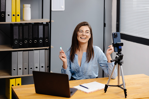 Videoconference And Training. Class of diverse students learning remotely online using web app. Female teacher providing lesson with laptop, recording video on cellphone on tripod.